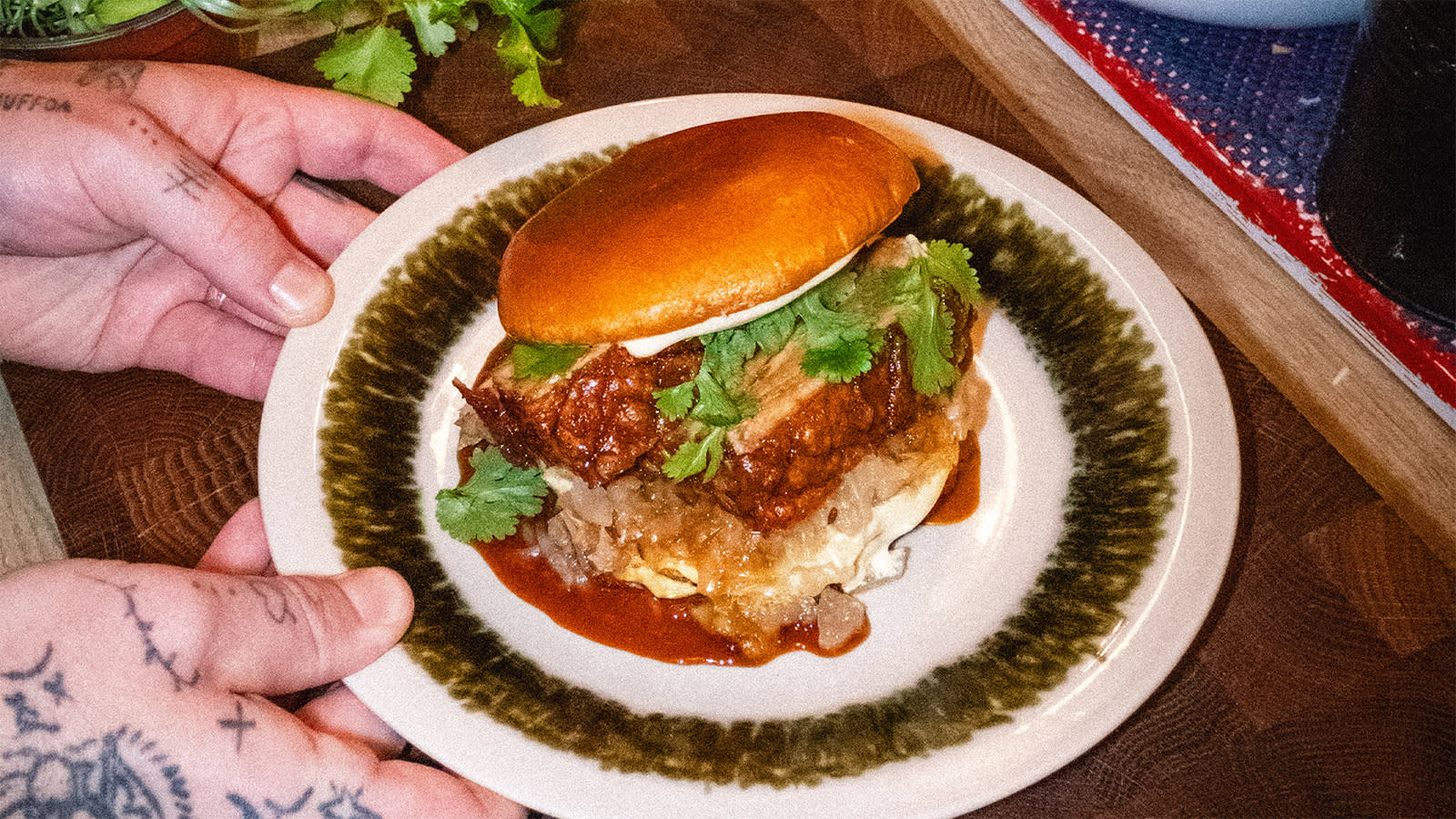 Ole Sirnes ribbeburger med surkål og teriyakisaus servert med miso-majo, briochebrød og koriander på hvit tallerken med mørkegrønt mønster.