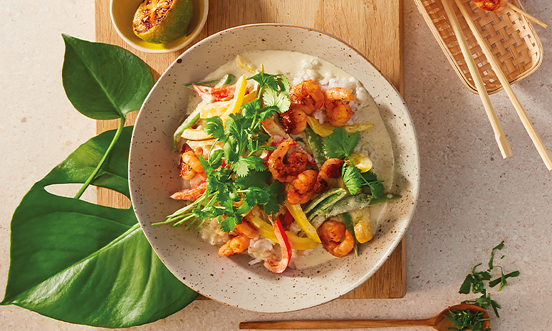 Green curry i prikkete hvit skål, med fargerike grønnsaker, fersk koriander og grillet scampi. Skålen står på en lys trefjøl med grillet lime på siden, og grønne blader til pynt.