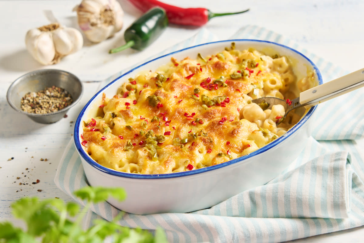 Hjemmelaget jalapeño mac'n cheese servert i hvit oval form med blå kant, på hvitt trebord med en liten skål pepper, hvitløk, jalapeño og rød chili i bakgrunnen.
