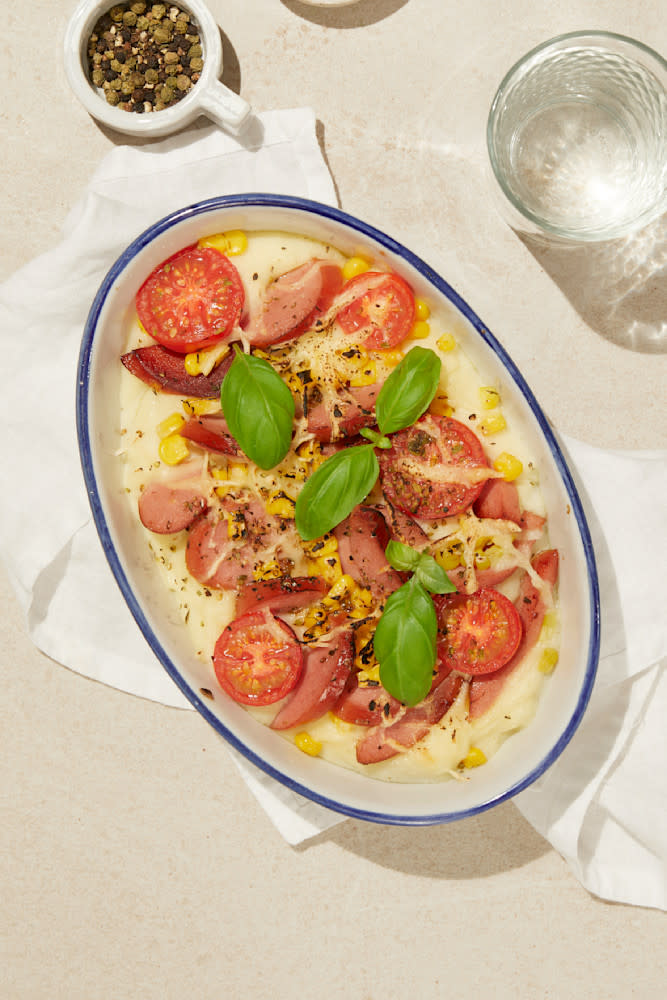 Pølsegrateng med potetmos, tomater og mais, servert i hvit ildfast form med blå kant. Glass med vann, og liten skål med hel pepper ved siden av.