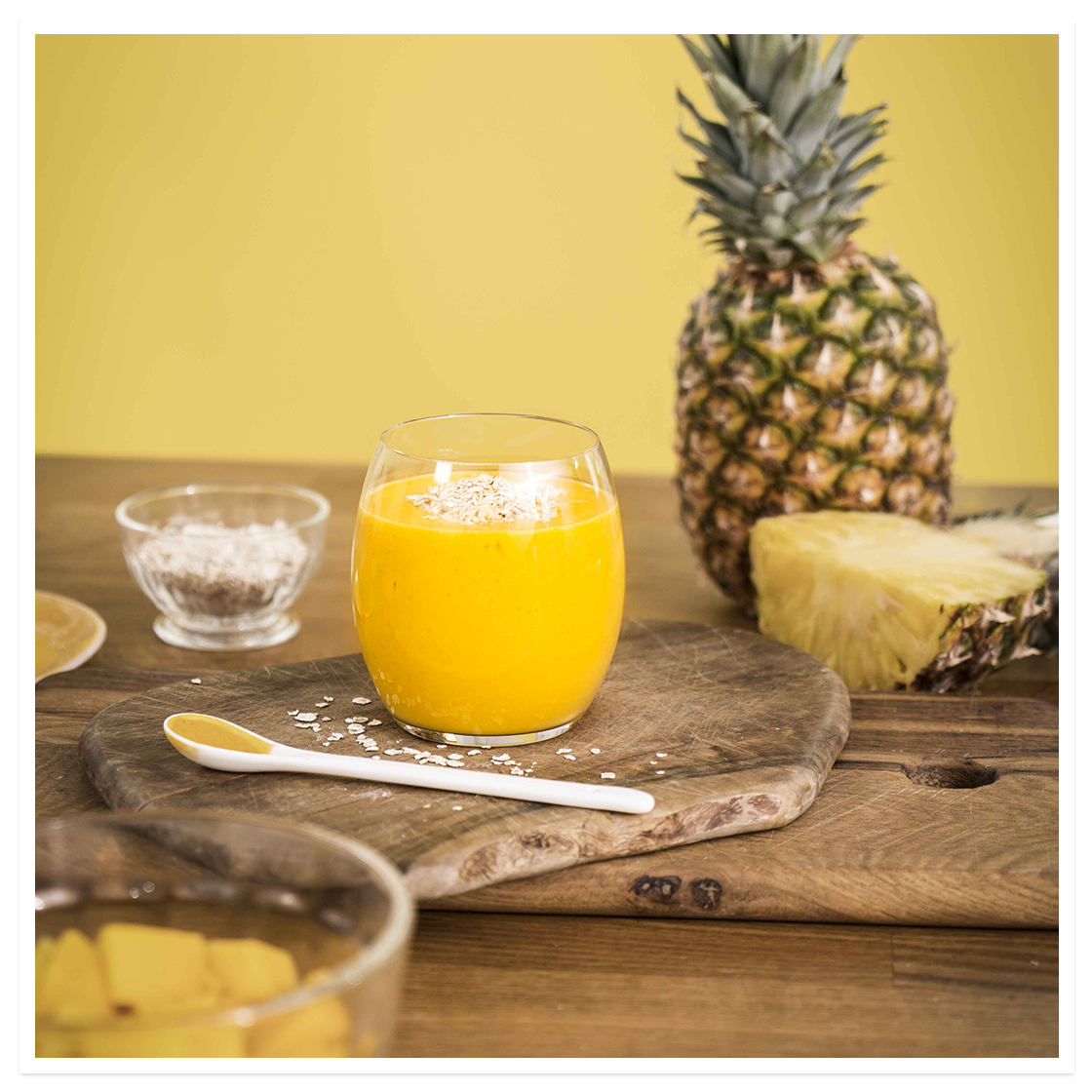 Solenergi -smoothie, gul smoothie med mango, ananas, gurkemeie og mandelmelk, i lite glass på trefjøl med fersk frukt rundt.