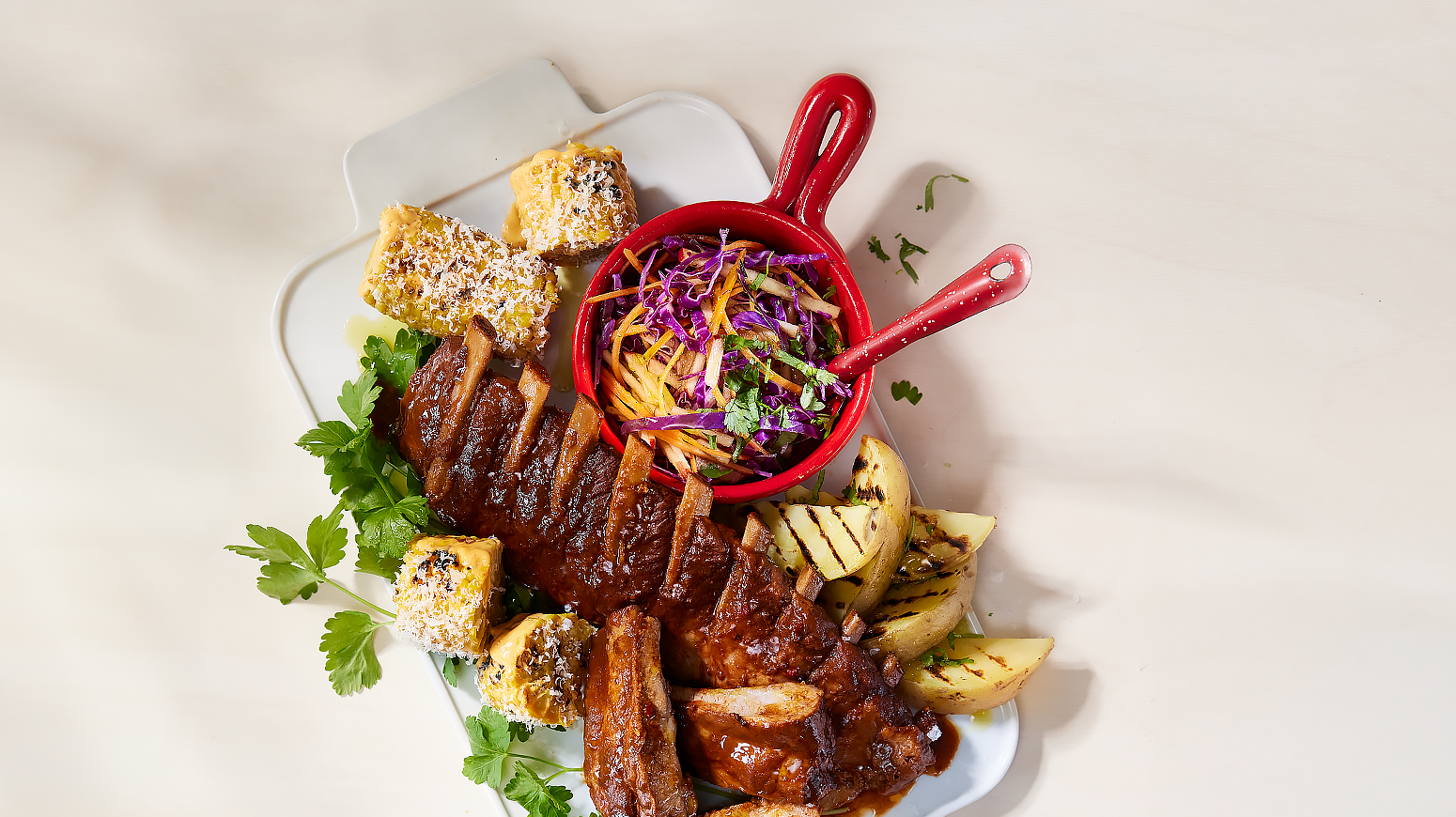 Spareribs med råkostsalat og grillet mais med parmesan, servert på hvitt fat med potetbåter og friske urter.