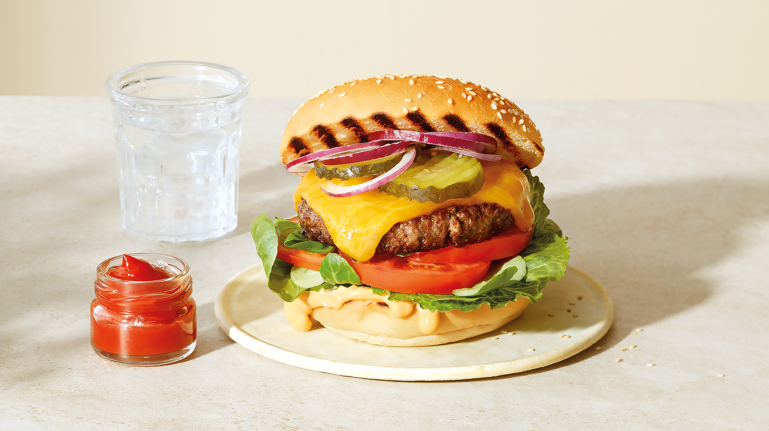 Klassisk cheeseburger med salat, tomat, dressing, rødløk og sylteagurk. Servert på lys tallerken med ketchup ved siden av.