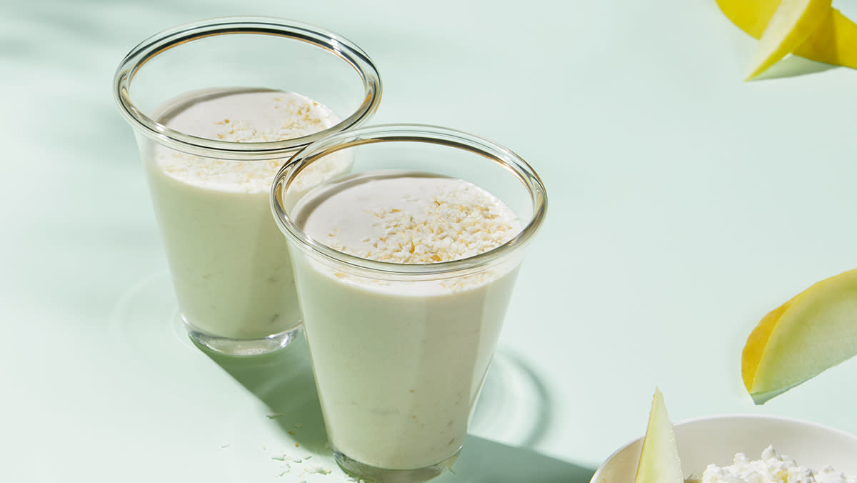 Sunn proteinsmoothie etter trening, servert i to glass på lys bakgrunn med sommerlig lys. Skiver av fersk honningmelon ved siden av.