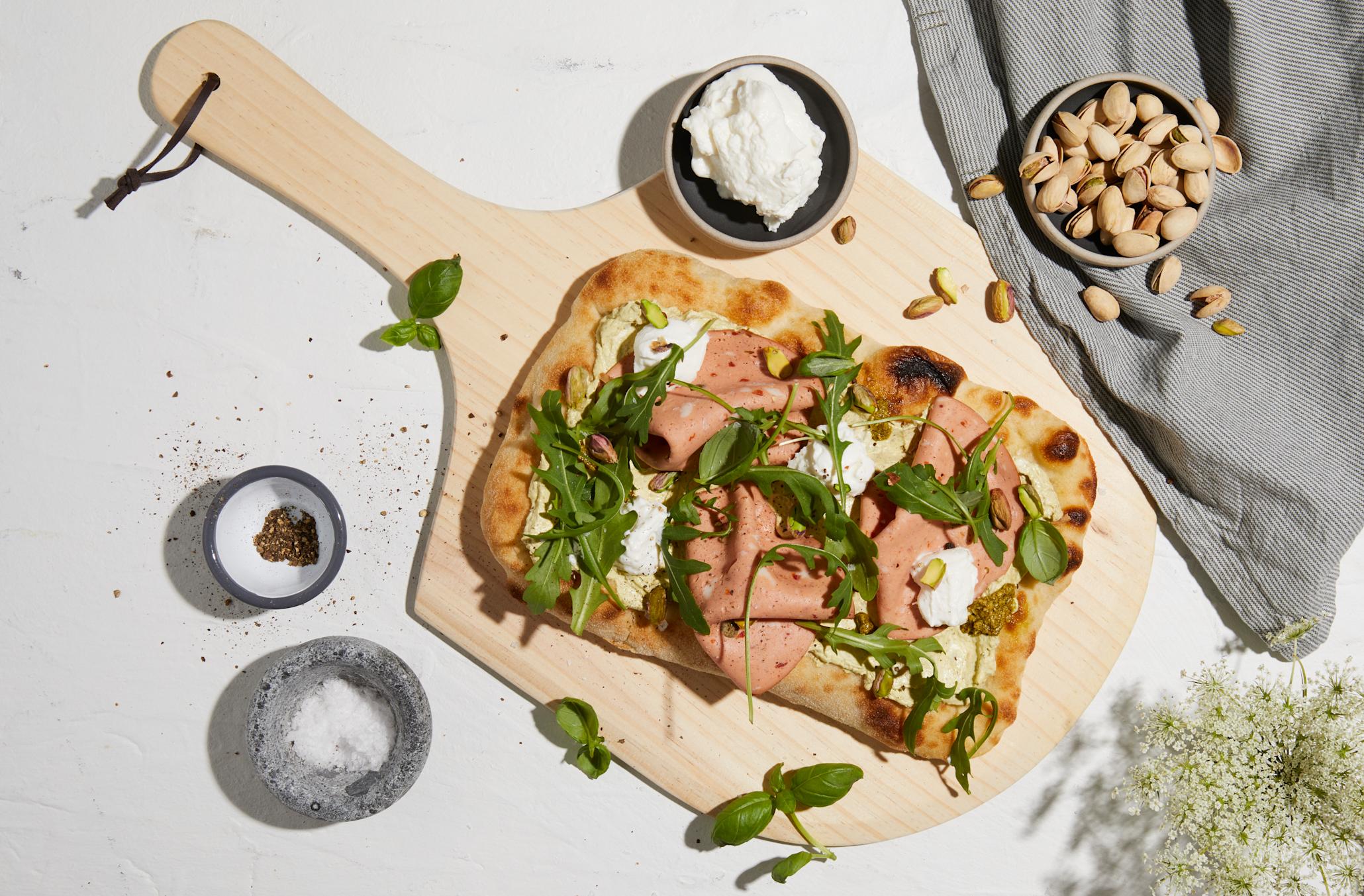 Grillet pinsa pizza med pestokrem, ricotta og mortadella servert på lys trefjøl med ruccola, pistasjenøtter og frisk basilikum.