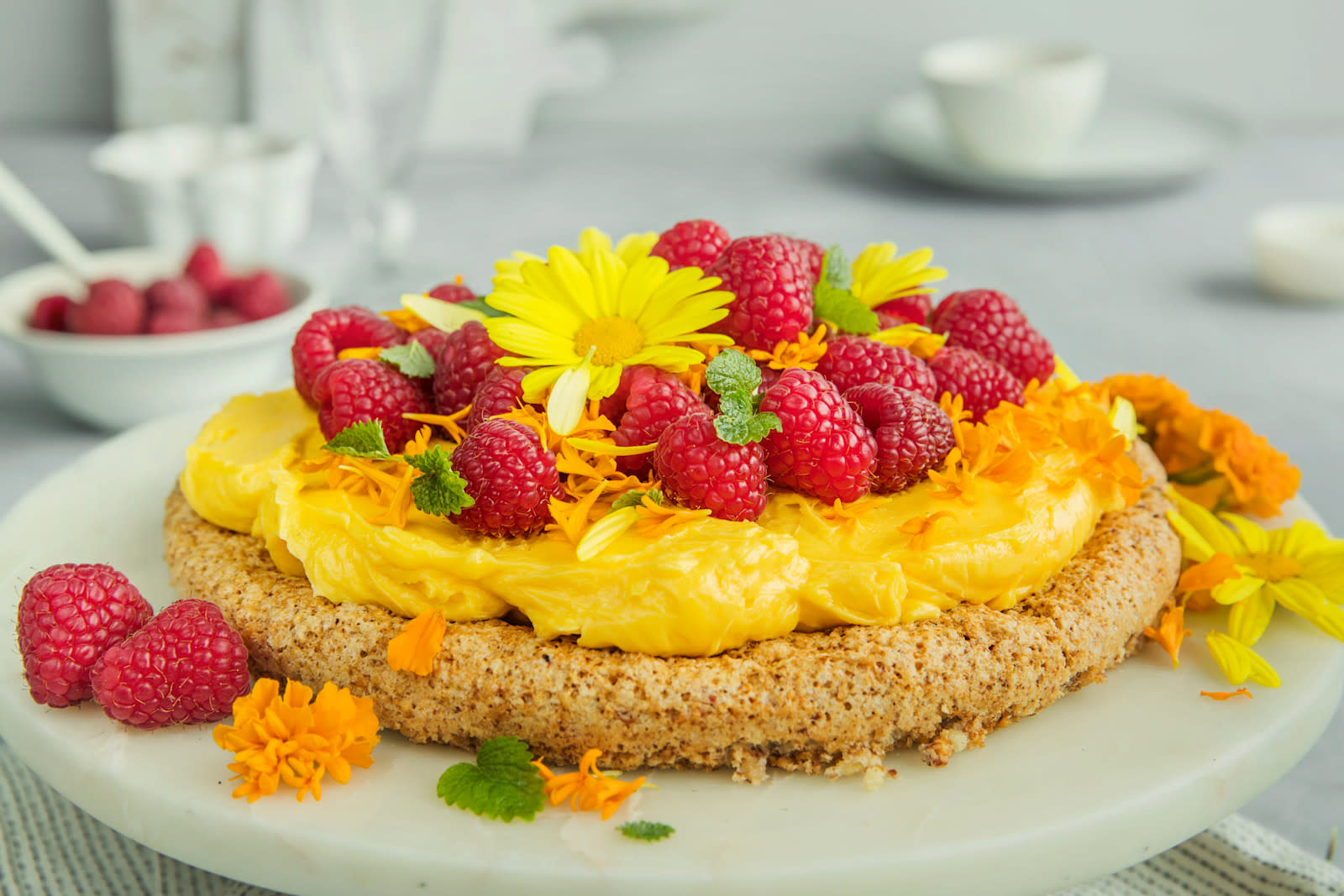 Suksessterte med friske bringebær, pyntet med sitronmelisse og friske blomster og servert på hvitt marmorfat på dekket kaffebord.