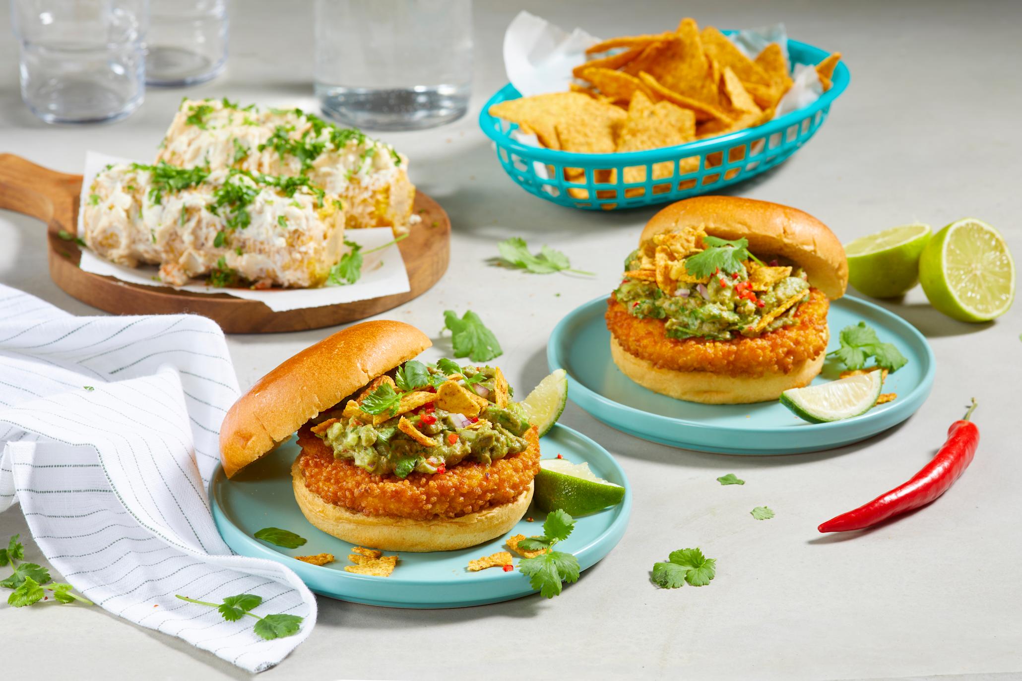 Crispy kyllingburger med spicy guacamole og meksikansk street corn servert på turkise asjetter på lyst bord med fersk rød chili og halve lime strødd rundt. Meksikansk street corn med fetaost og koriander, og en turkis kurv med tortilla chips i bakgrunnen.