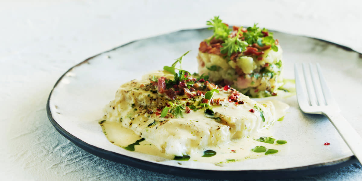 Lutefisk med julekrydder, sennepsskum, poteter og bacon servert på rustikk hvit tallerken med friske urter over og gaffel ved siden av.