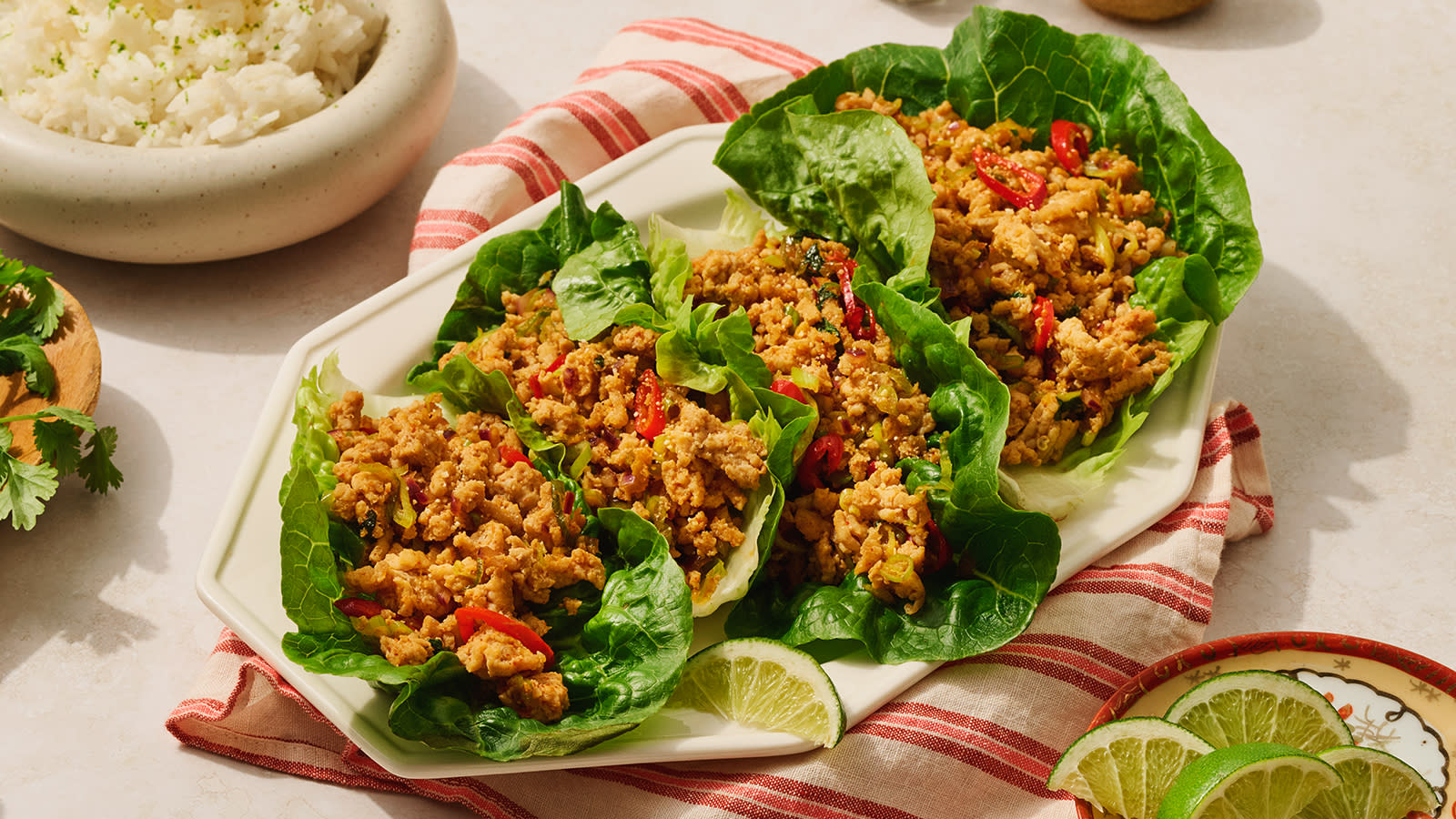 Thai laab gai, krydret kyllingkjøttdeig servert i salatblader, på hvit asjett på rødstripet kjøkkenhåndkle. Skal med ris i bakgrunnen.