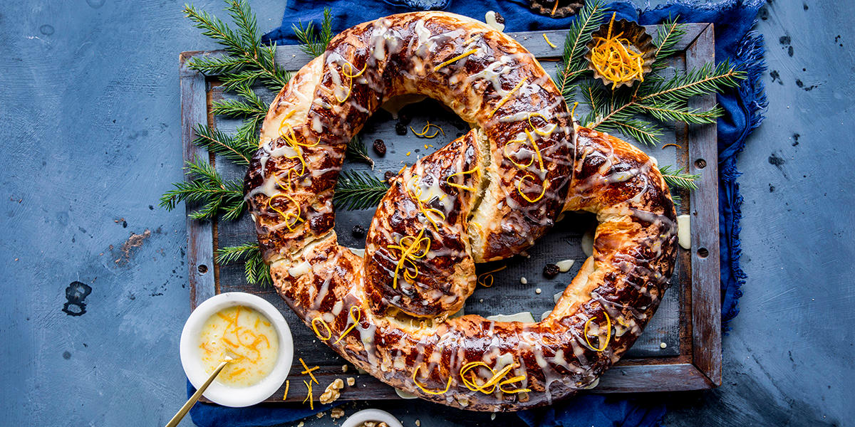 Kringle med marsipan og appelsinglasur, servert på trefjøl med granbar, revet appelsinskall, og en skål appelsinglasur ved siden av. Mørkeblått kjøkkenhåndkle på kanten.
