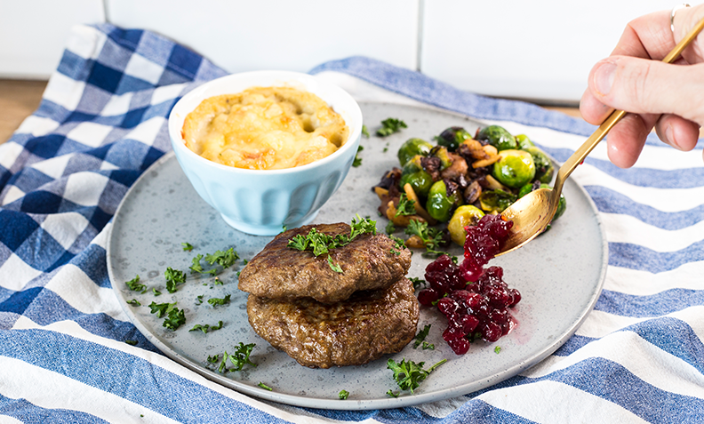 Karbonader på grå middagstallerken, med makaronigrateng i liten skål, og rosenkål med bacon ved siden av. Blårutet kjøkkenhåndkle i bakgrunnen, og hånd som serverer tyttebærsyltetøy.