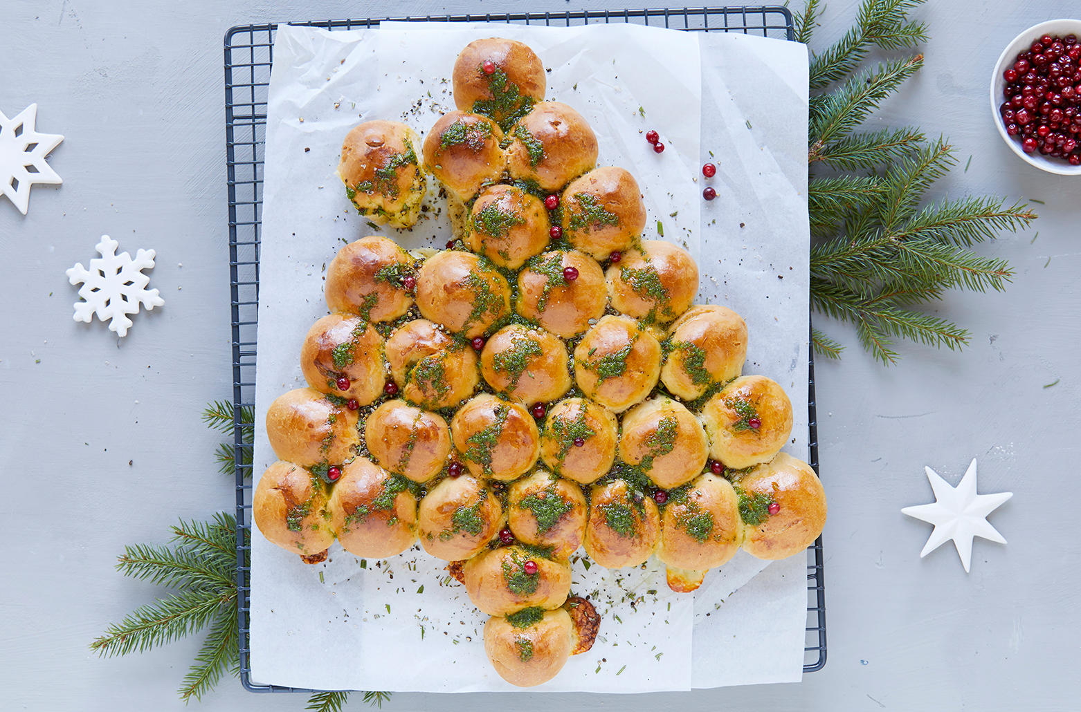 Fylte juletrerundstykker med mozzarella, satt sammen til et juletre, og toppet med grønn urteolje. Juletrerundstykkene er servert på svart kjølerist med hvitt bakepapir, og pyntet med ferske tyttebær. Granbar og hvite julestjerner i bakgrunnen.