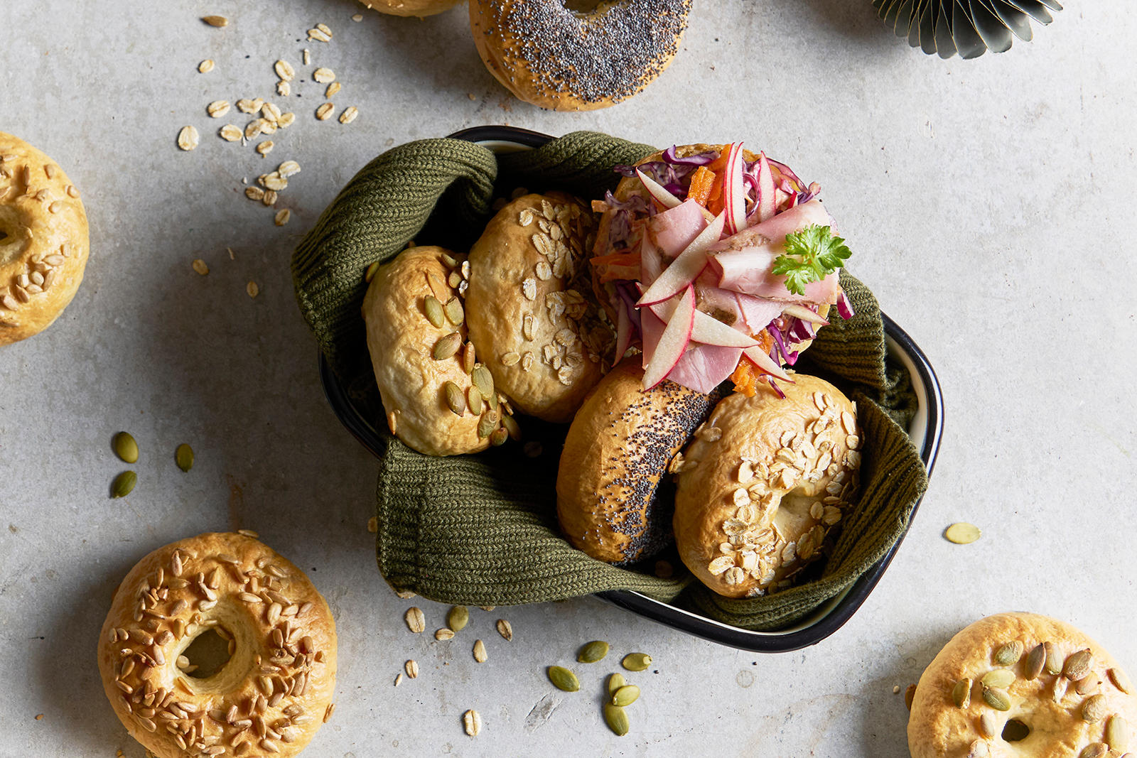 Hjemmelagede bagels med frø og kjerner, servert i kurv med grønt kjøkkenhåndkle. En bagel er servert med eple, ribberull og rødkål.