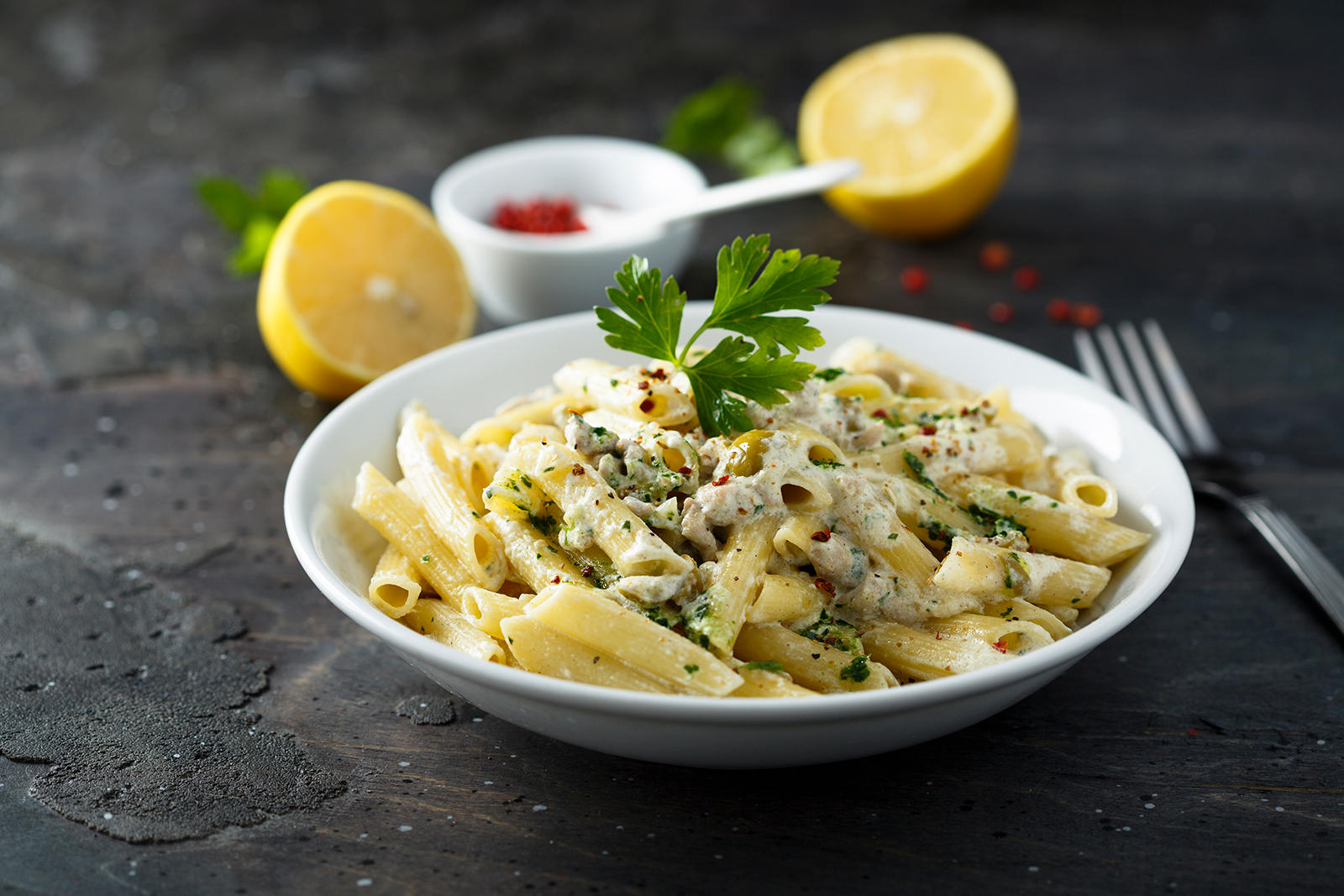 Pasta al limone med penne pasta, servert i hvit skål på mørk bakgrunn. Halve friske sitroner, og grønne urter i bakgrunnen.