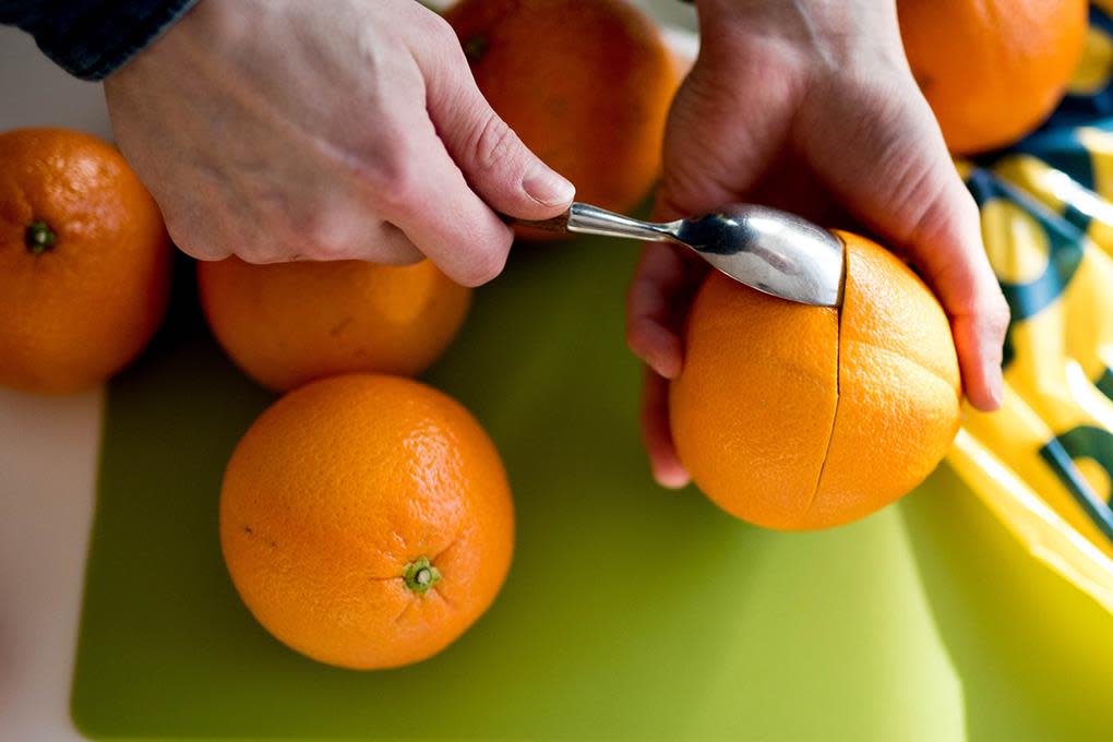 To hender holder i en appelsin, og skreller den med en skje. Grønt skjærebrett som underlag, og flere appelsiner rundt.