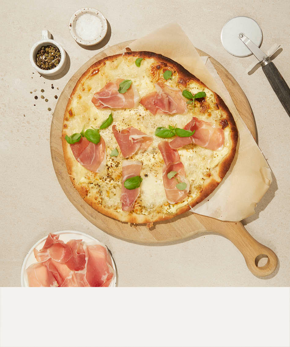 Pizza med parmaskinke og basilikum på fjøl på høyre og en beige stripe nederst